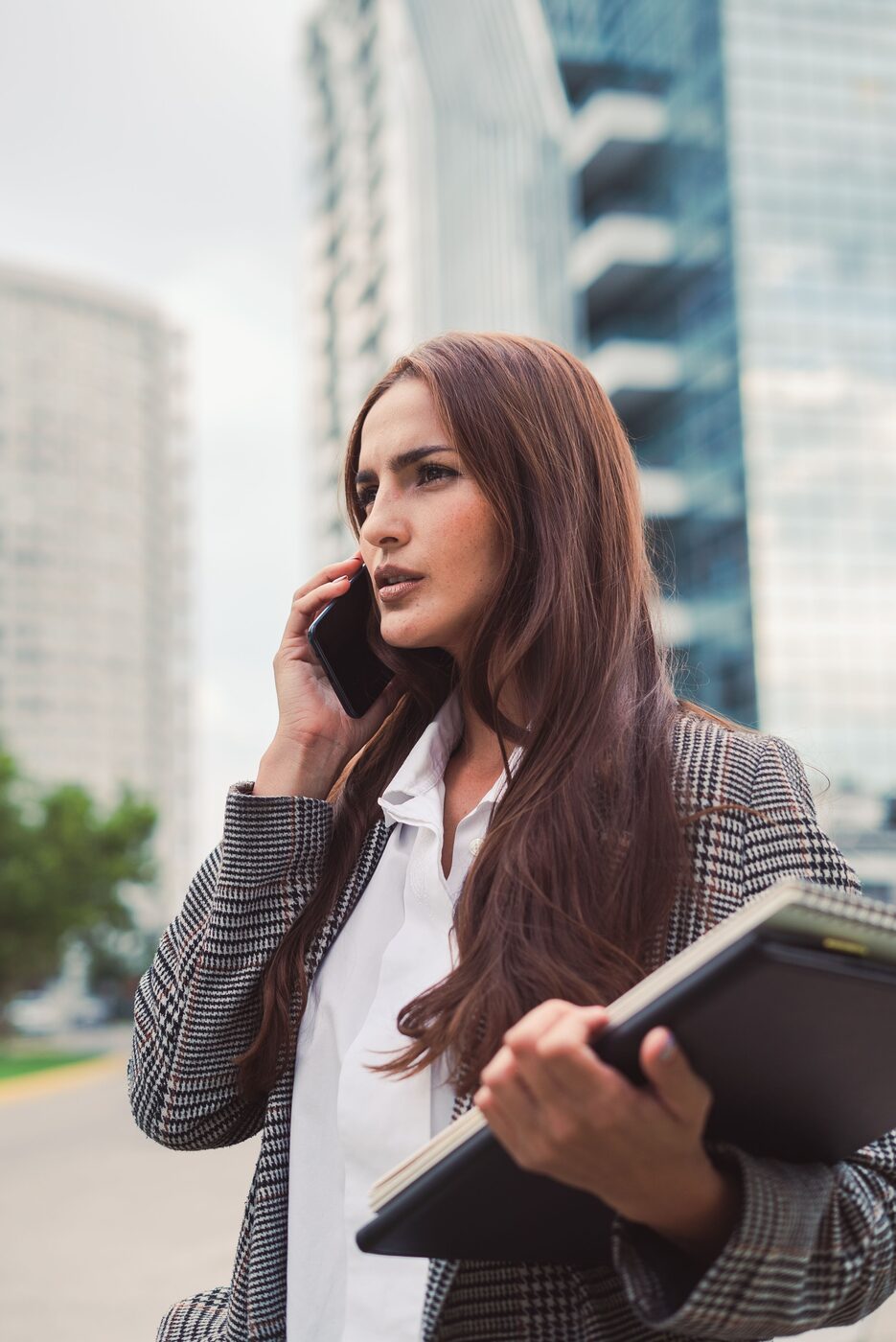 Kobieta w miejskiej zabudowie rozmawia przez telefon, trzymając dokumenty